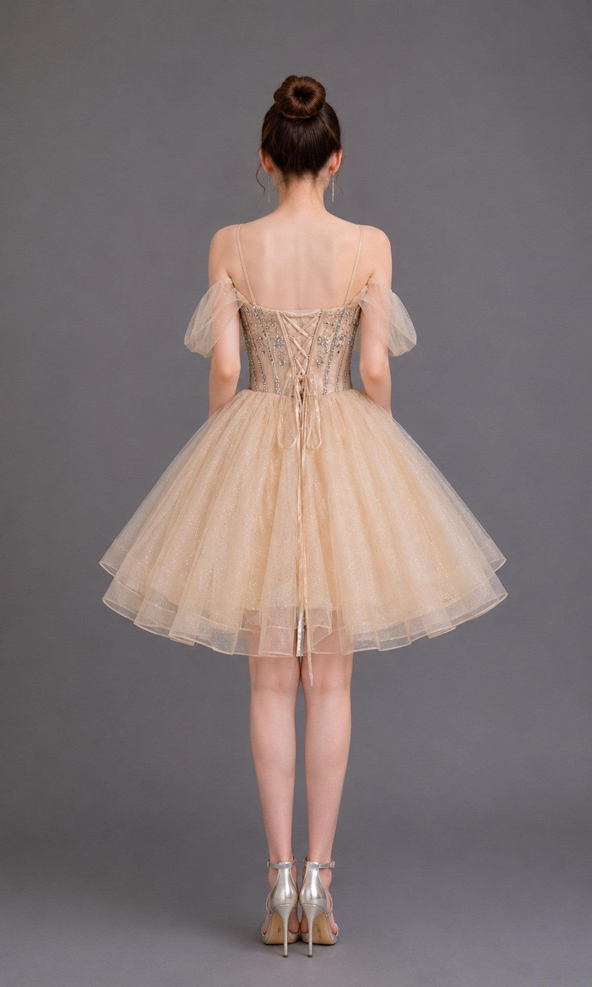 A woman models the Short Homecoming Dress: Dancing Queen 3438, featuring off-the-shoulder straps, a lace-up back, and a sparkly tulle skirt, as she stands against a plain gray background.